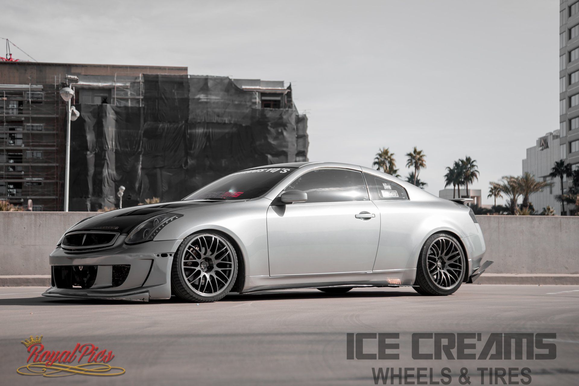 Lowered silver sports coupe parked on concrete with city buildings in the background.