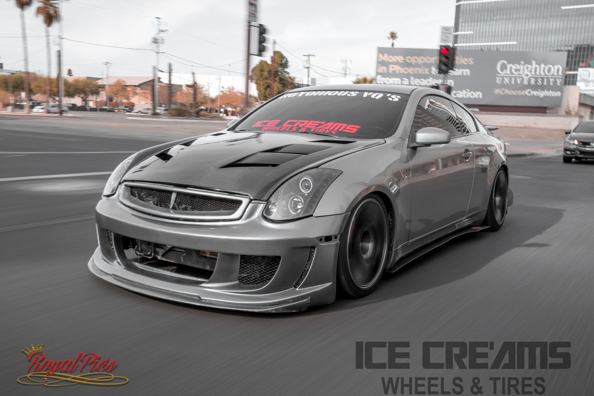 Low gray sports coupe with wide body kit parked on a street, tire shop branding visible in background