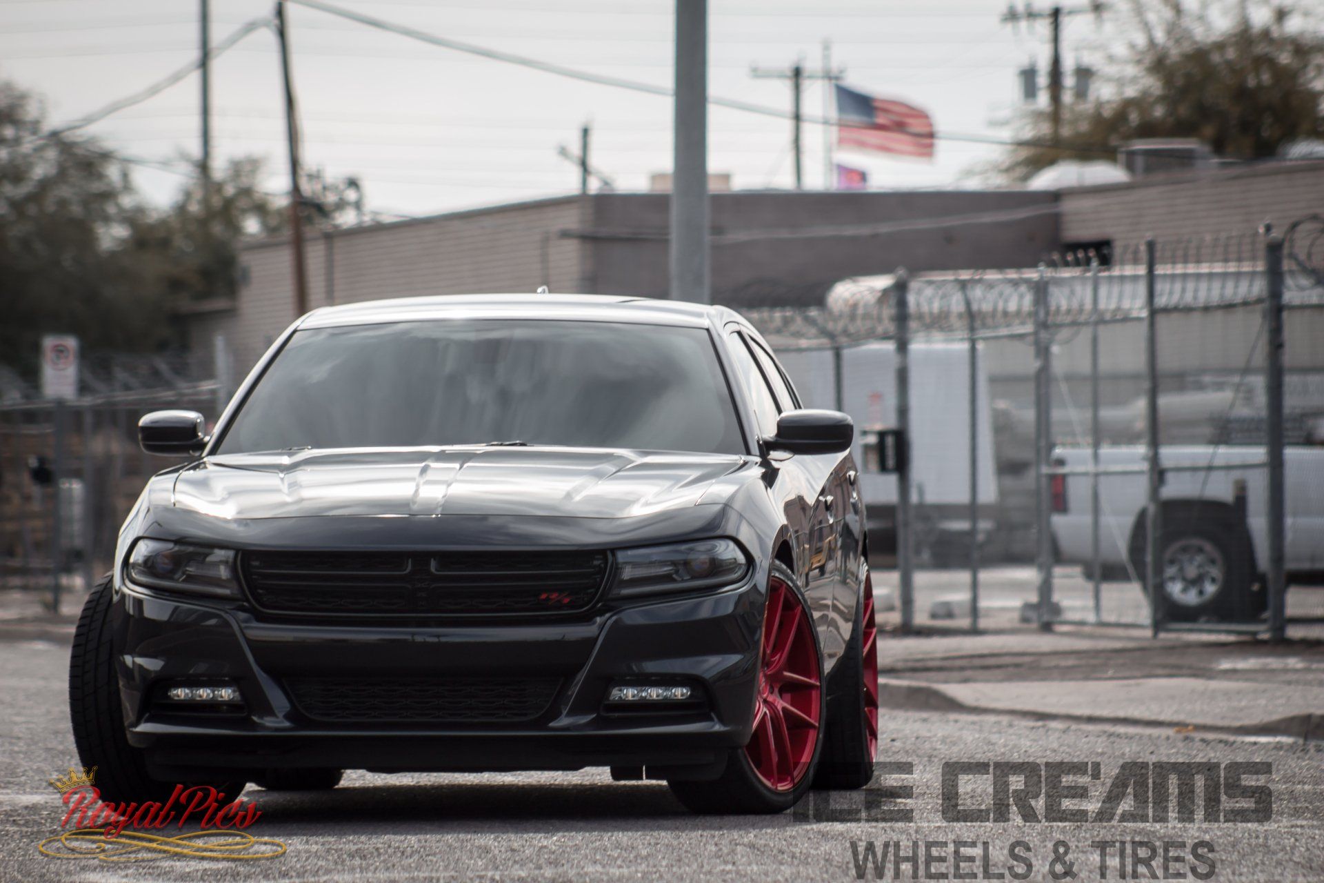 Black Dodge Charger with red rims parked on a street near a chain-link fence and industrial buildings