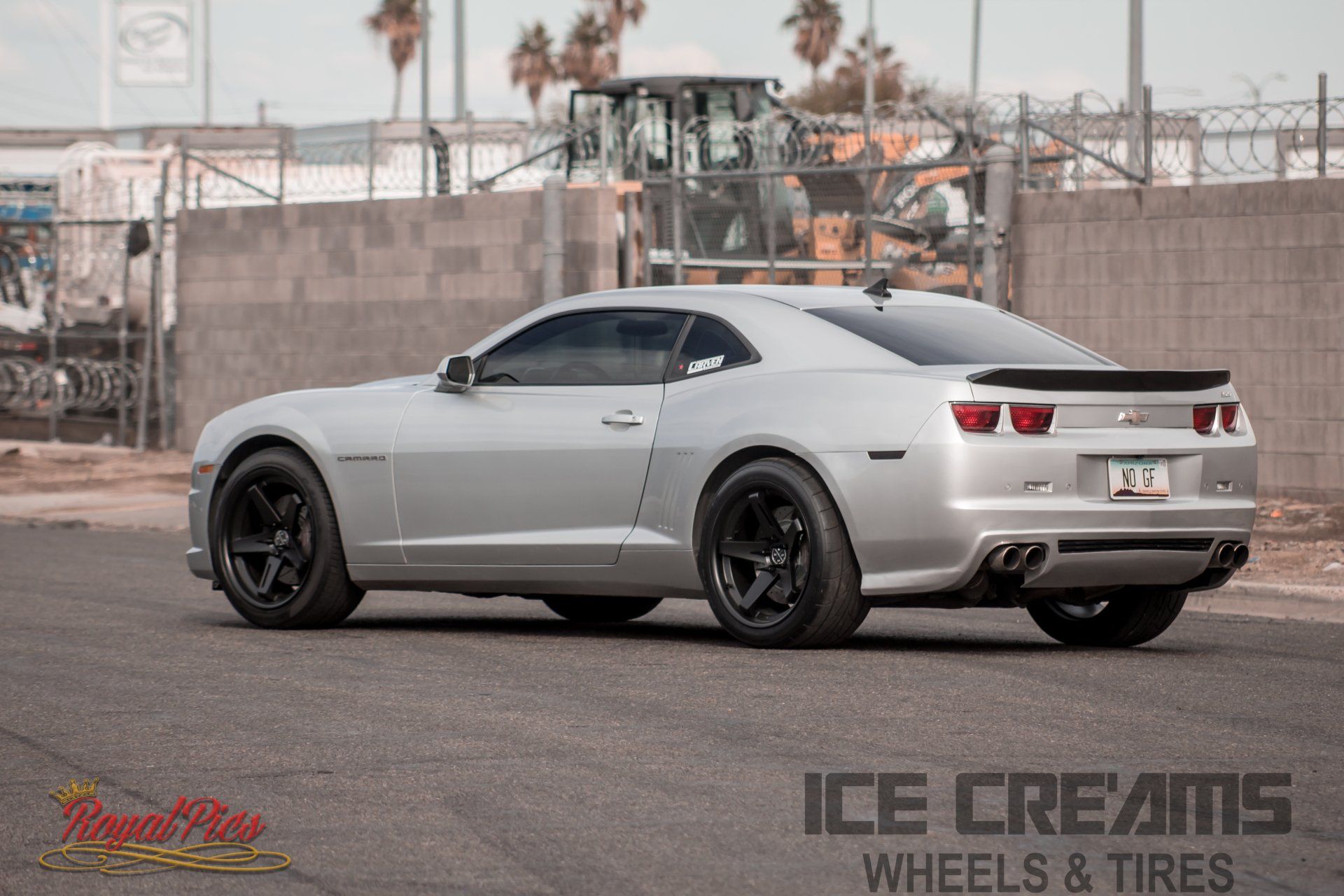 Silver Chevrolet Camaro with black wheels parked outdoors, side view against a fenced industrial background.