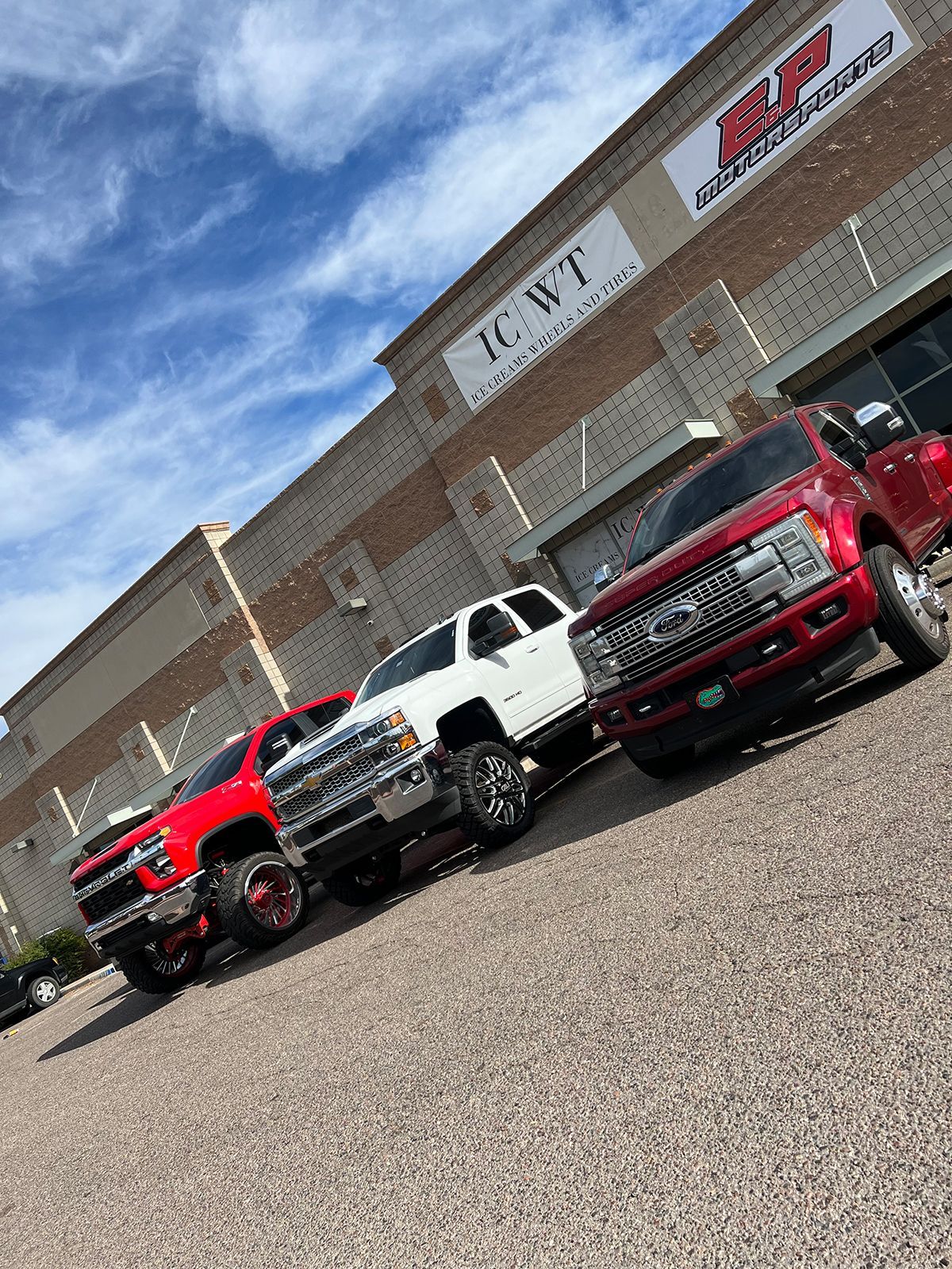 Three lifted trucks parked outside a brick auto dealership under a blue sky.