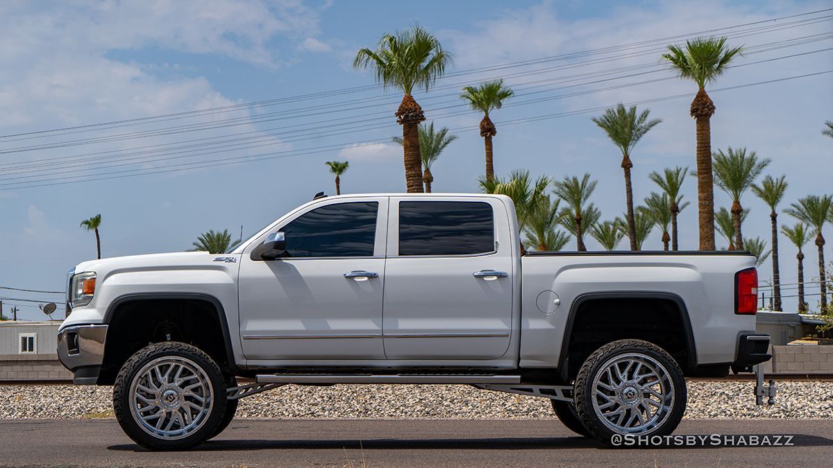 Silver lifted pickup truck parked on gravel with palm trees in the background.