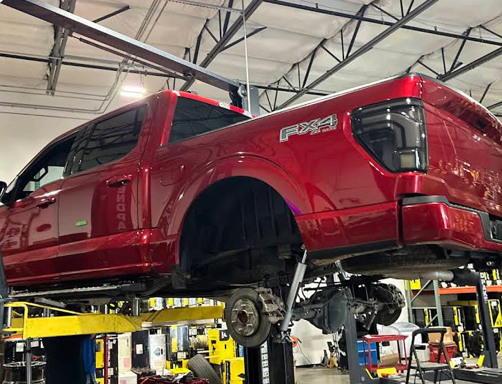 Red pickup truck lifted on a shop hoist inside an auto repair garage, wheels removed.