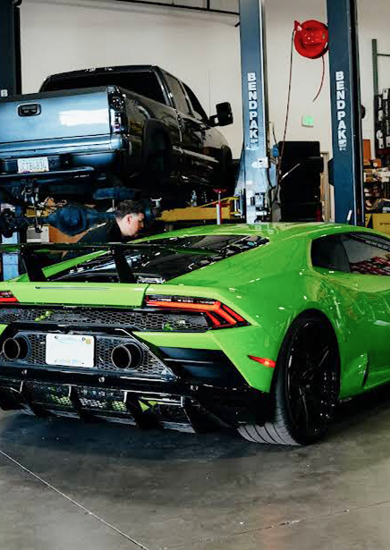Green Lamborghini in auto shop with a mechanic working under a raised black pickup truck.