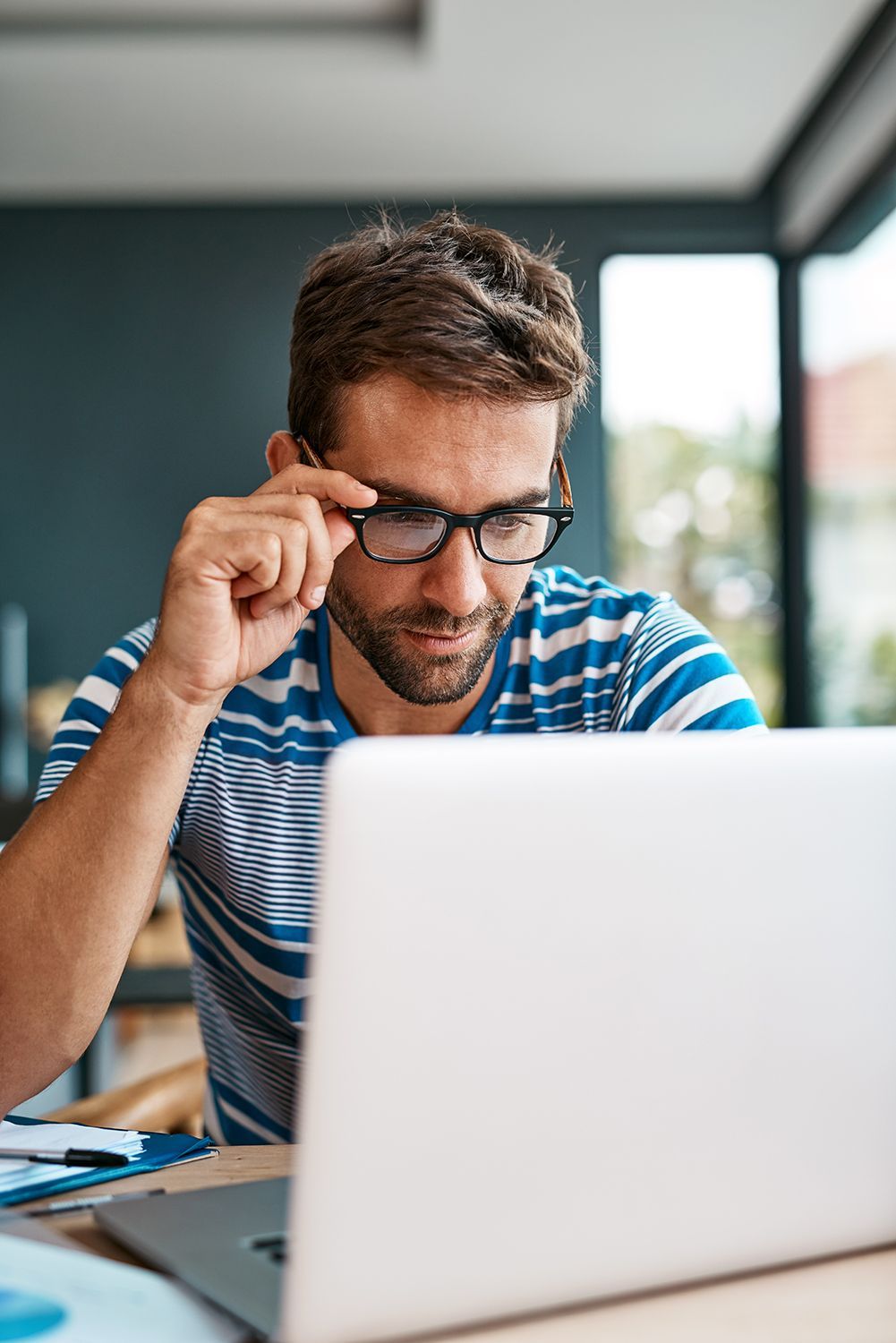 Man adjusting glasses while working on a laptop at a desk near a bright window