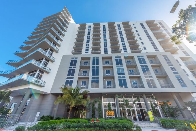 Modern high-rise building with balconies and a glass entrance, under a bright blue sky.