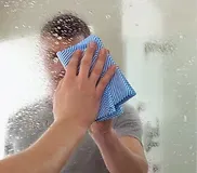 A hand wipes a foggy bathroom mirror with a blue cloth, clearing condensation.