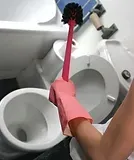 Person in pink gloves cleaning a white toilet with a red brush in a bathroom