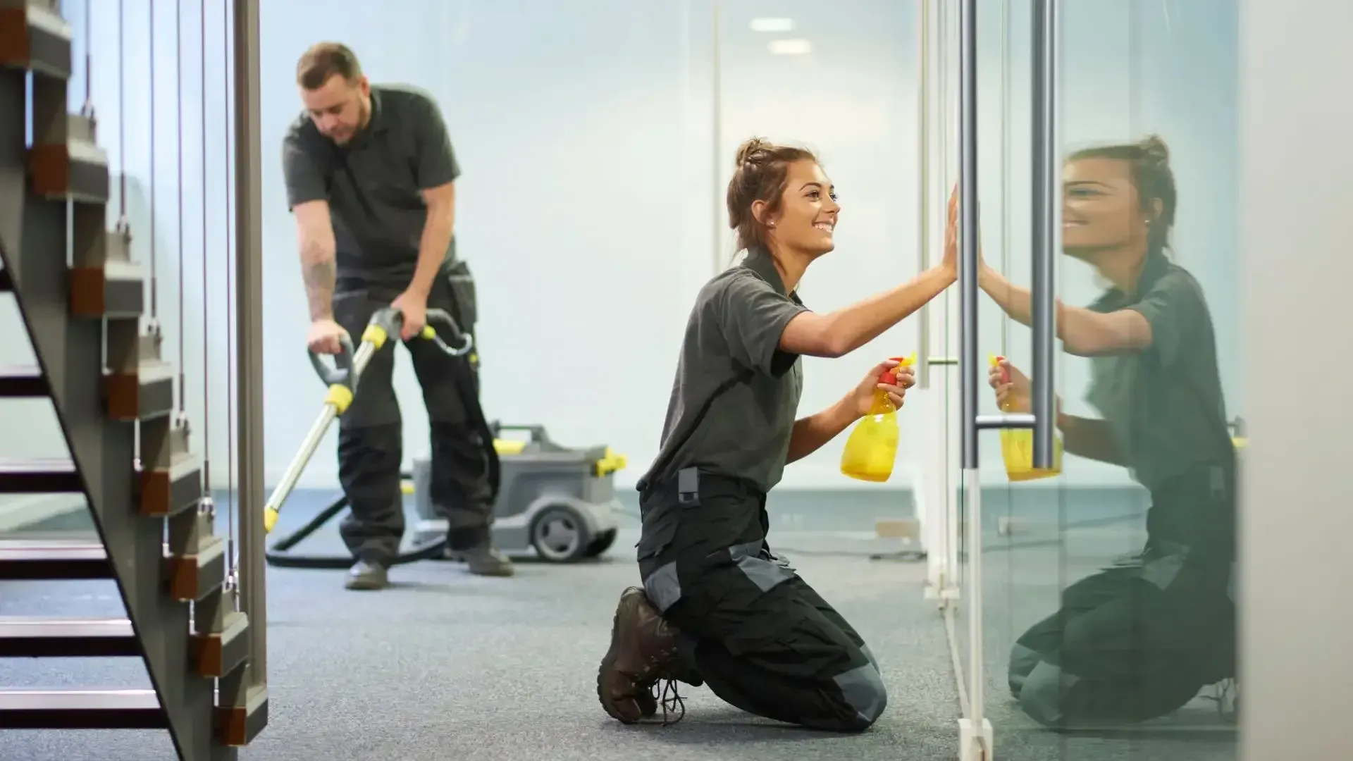 Two cleaners in an office, one vacuuming a carpet and one kneeling to wipe a glass door.