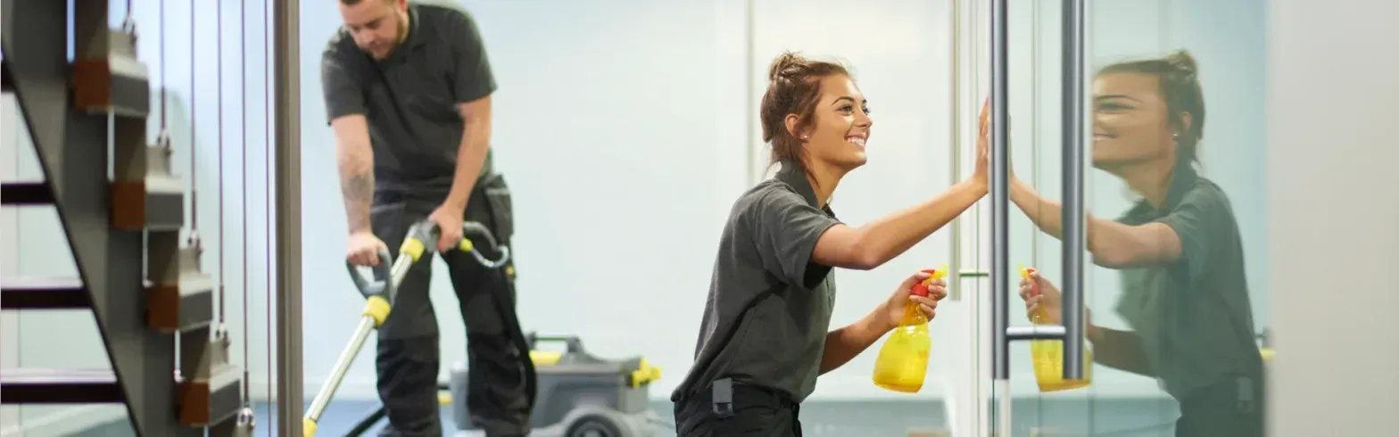 Woman wiping a glass door while a man vacuums the floor in a modern office lobby