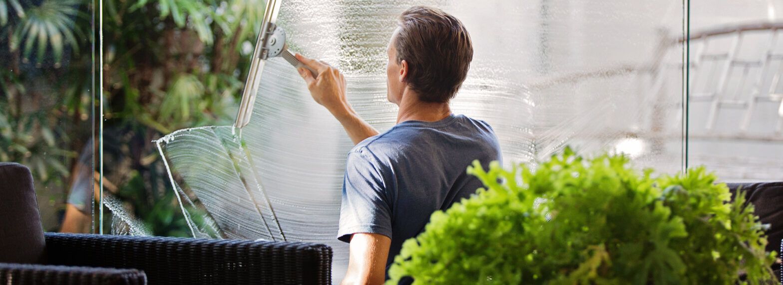 Person watering plants in a greenhouse, with lush green foliage in the foreground