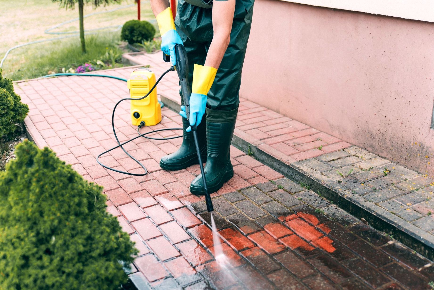 Person pressure-washes a brick walkway beside a pink wall, cleaning a wet section. Person pressure-washes a brick walkway beside a pink wall, cleaning a wet section.
