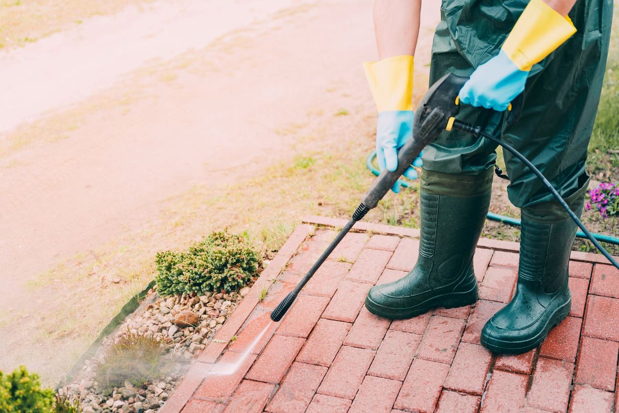 Worker pressure-washing brick pavement beside a curb, wearing black boots and gloves Worker pressure-washing brick pavement beside a curb, wearing black boots and gloves
