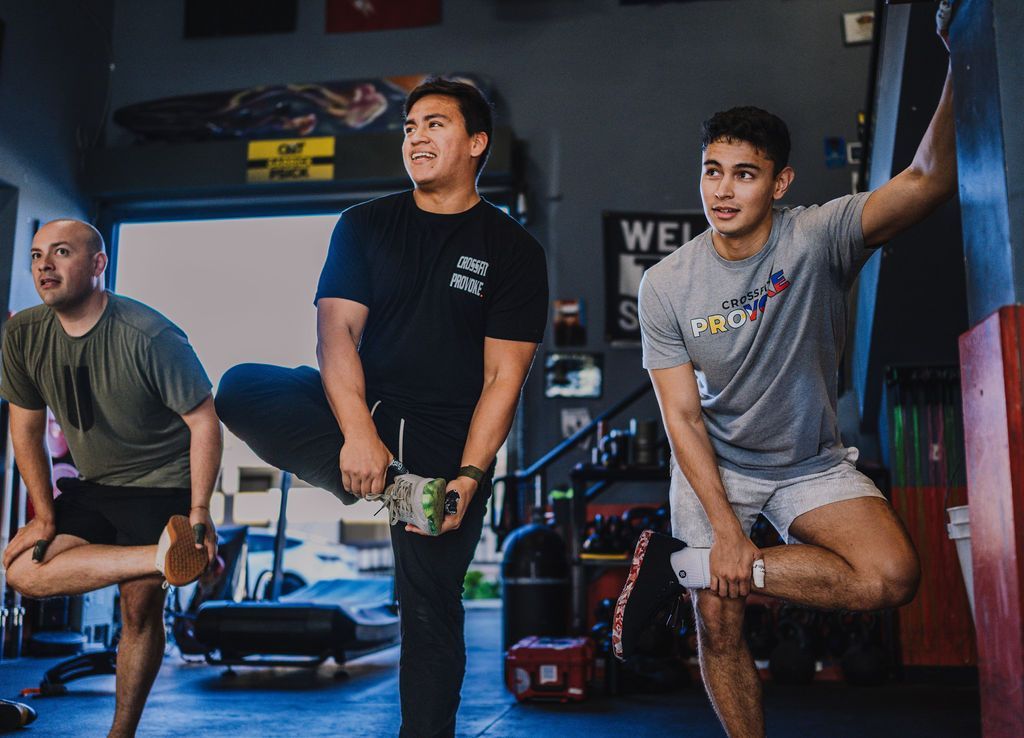 Three men stretching in a gym: lifting legs, smiling. Interior with equipment and open doorway.