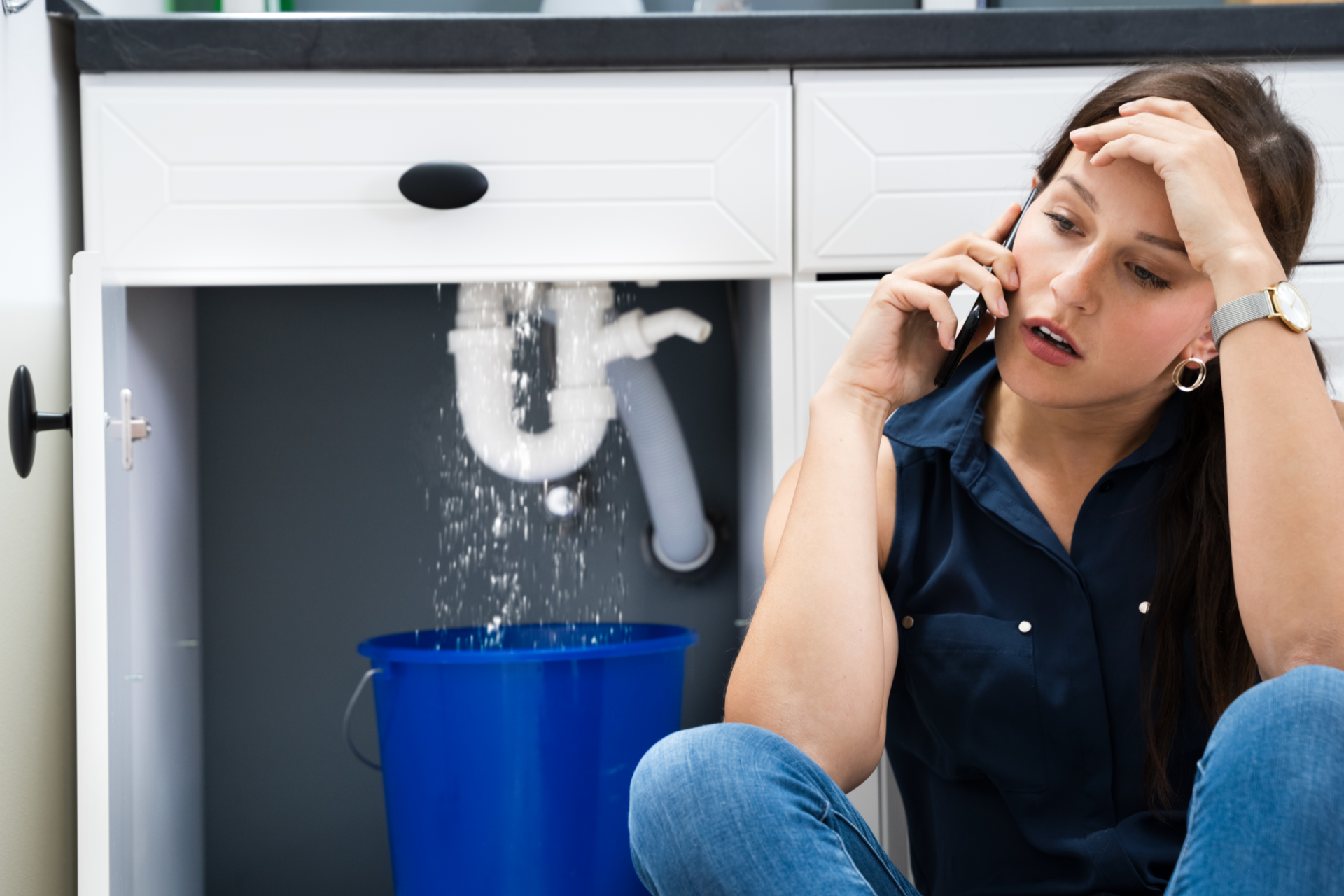 Woman on phone beside a leaking sink, with water dripping into a blue bucket under the cabinet.