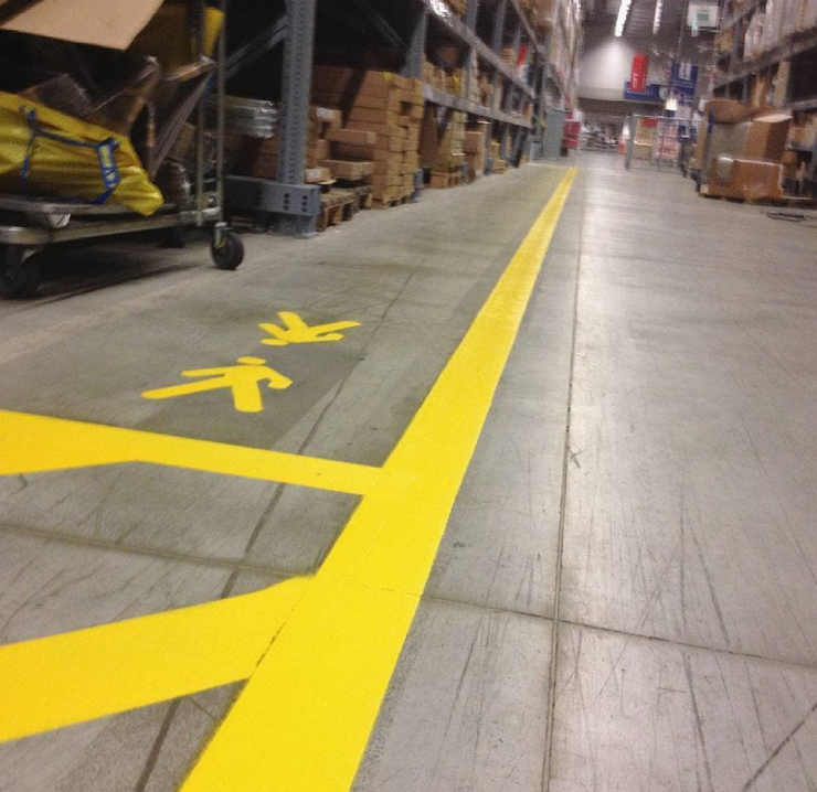 Corsia di magazzino con una linea di sicurezza giallo brillante sul pavimento, accanto a scaffali carichi di merce e carrelli.