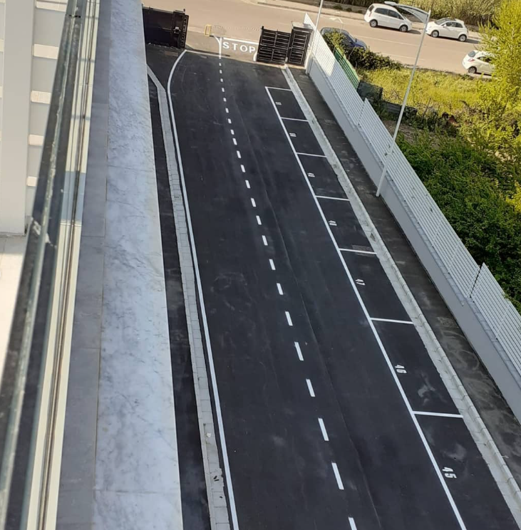 Vista dall’alto di una stretta corsia carrabile con segnaletica bianca, delimitata da muri e con auto parcheggiate in lontananza