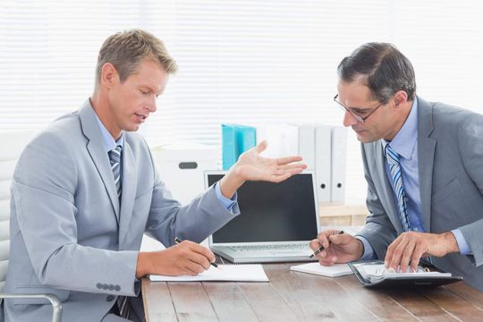 two men sitting at a table working on a laptop