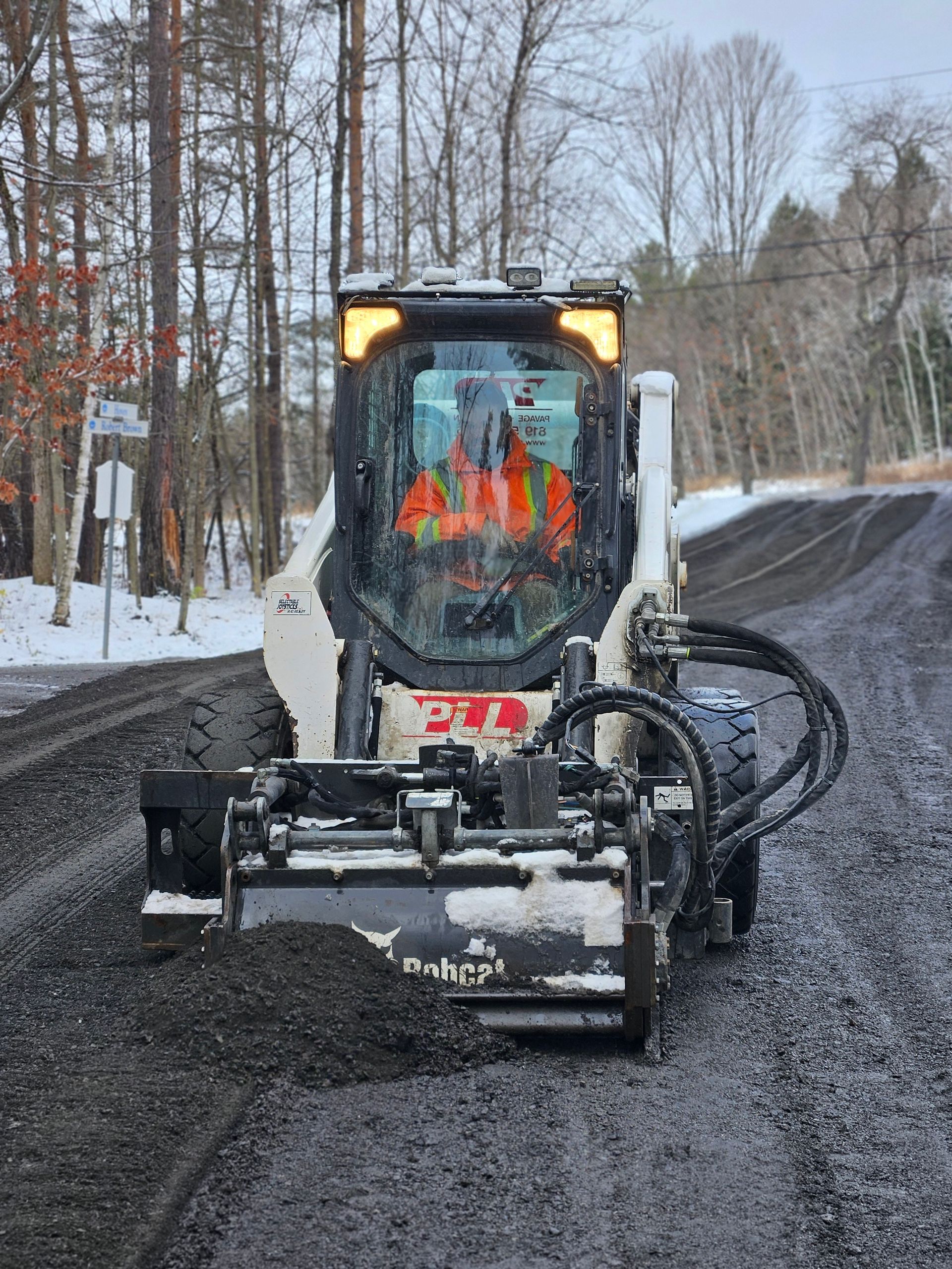 Une déneigeuse dégage une route enneigée, avec un conducteur en gilet orange dans sa cabine, dans une rue forestière en hiver.