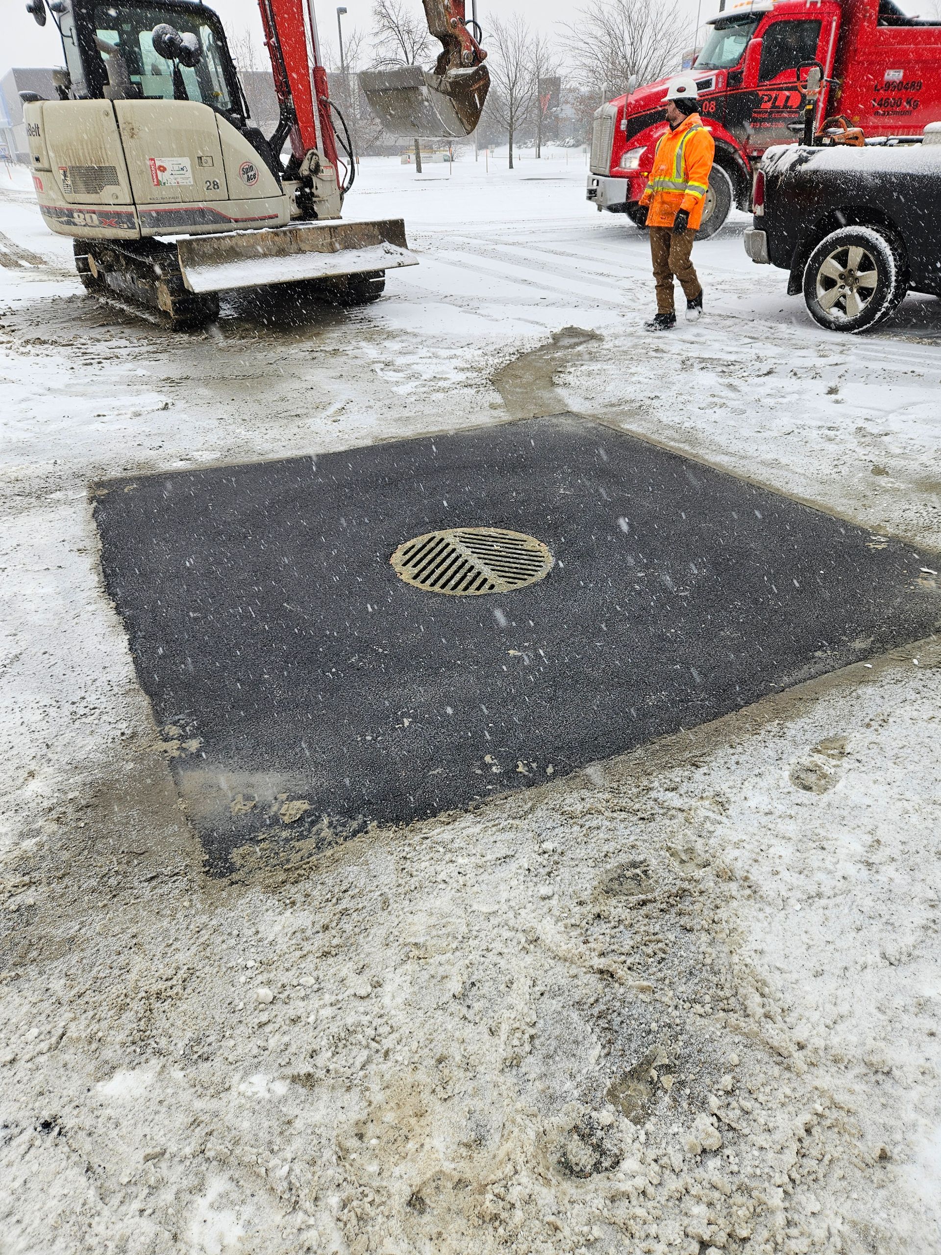 Rue enneigée avec une plaque d'égout réparée, un camion de chantier et un ouvrier en tenue de sécurité orange.