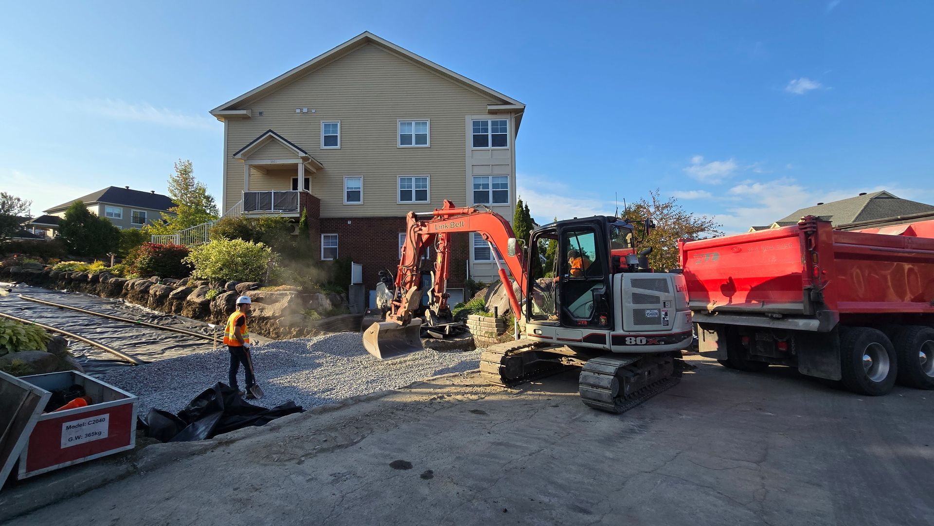 Des ouvriers du bâtiment et une pelleteuse démolissent un mur de pierre à côté d'une maison beige et d'un camion-benne rouge.