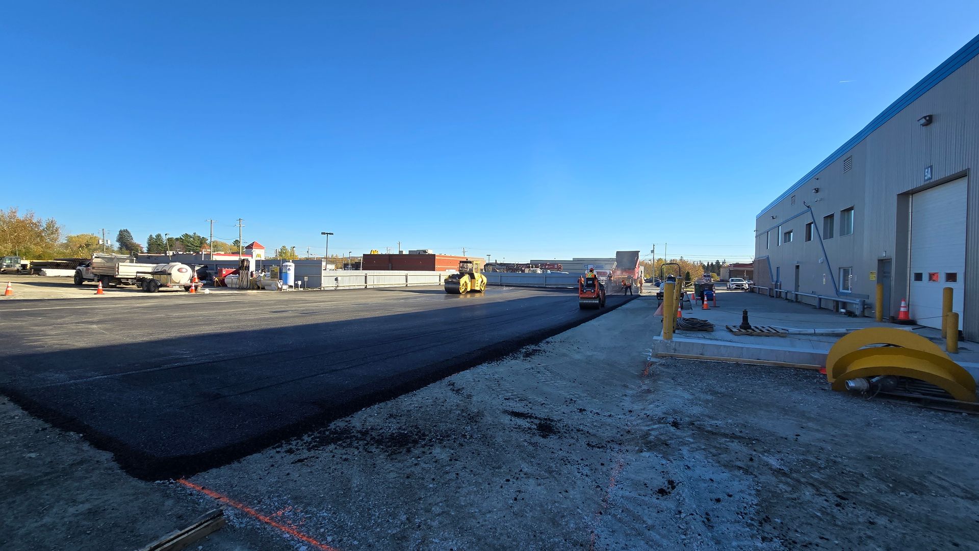 Travaux de pavage de route à côté d'un entrepôt, avec de l'asphalte noir neuf, des engins de chantier et des ouvriers.