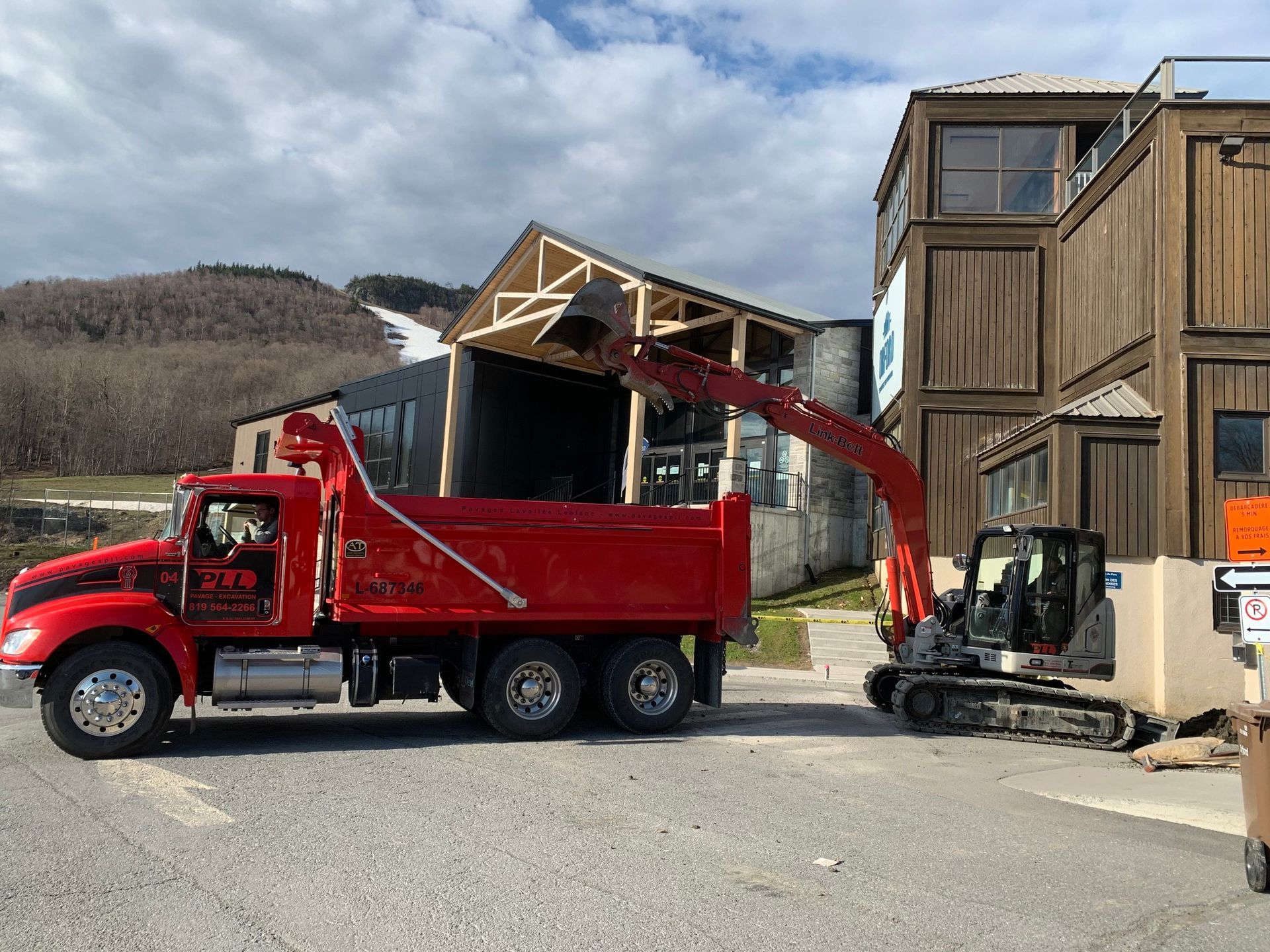 Un camion-benne rouge charge des débris à côté d'une petite pelle mécanique sur un chantier de construction.