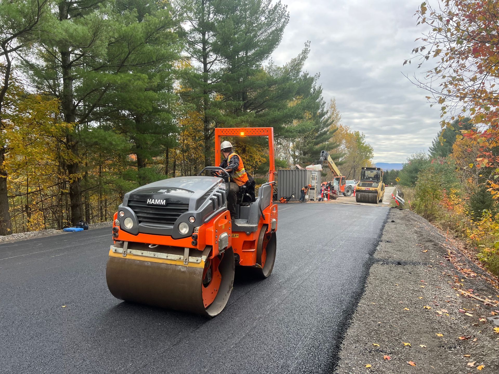 Un rouleau compresseur orange asphalte une route rurale, des ouvriers derrière lui.