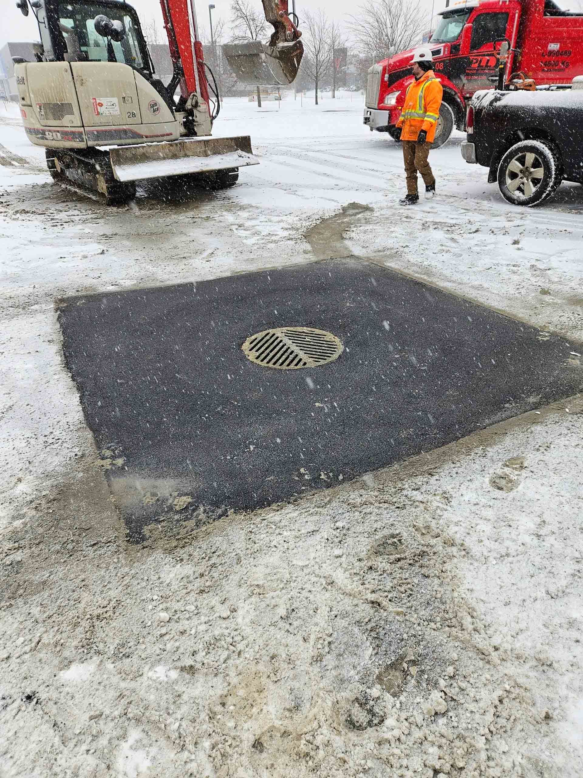 Scène de travaux routiers sous la neige : un ouvrier se trouve près d’un camion et d’une plaque d’égout fraîchement réparée.