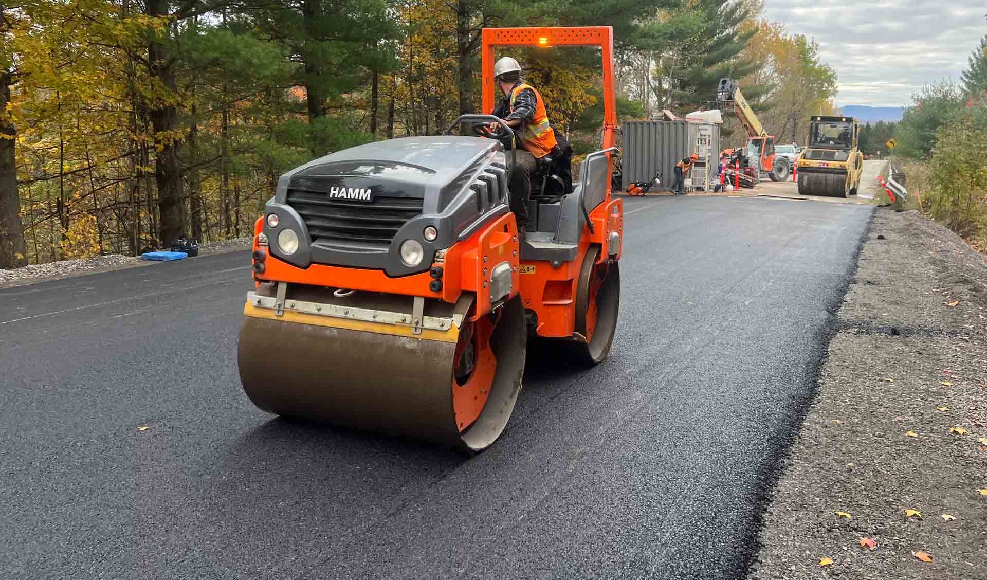 Rouleau compresseur orange sur une route nouvellement asphaltée, avec des ouvriers et des engins de chantier en arrière-plan.