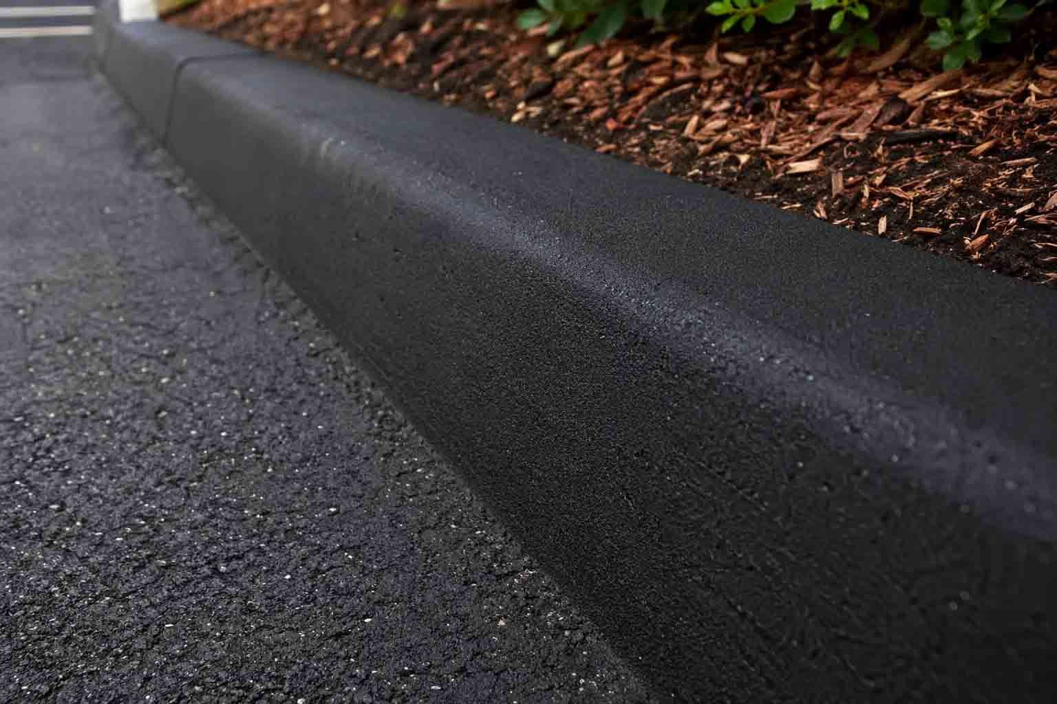 Bordure incurvée en béton foncé le long d'une allée asphaltée, bordée de paillis et de plantes.