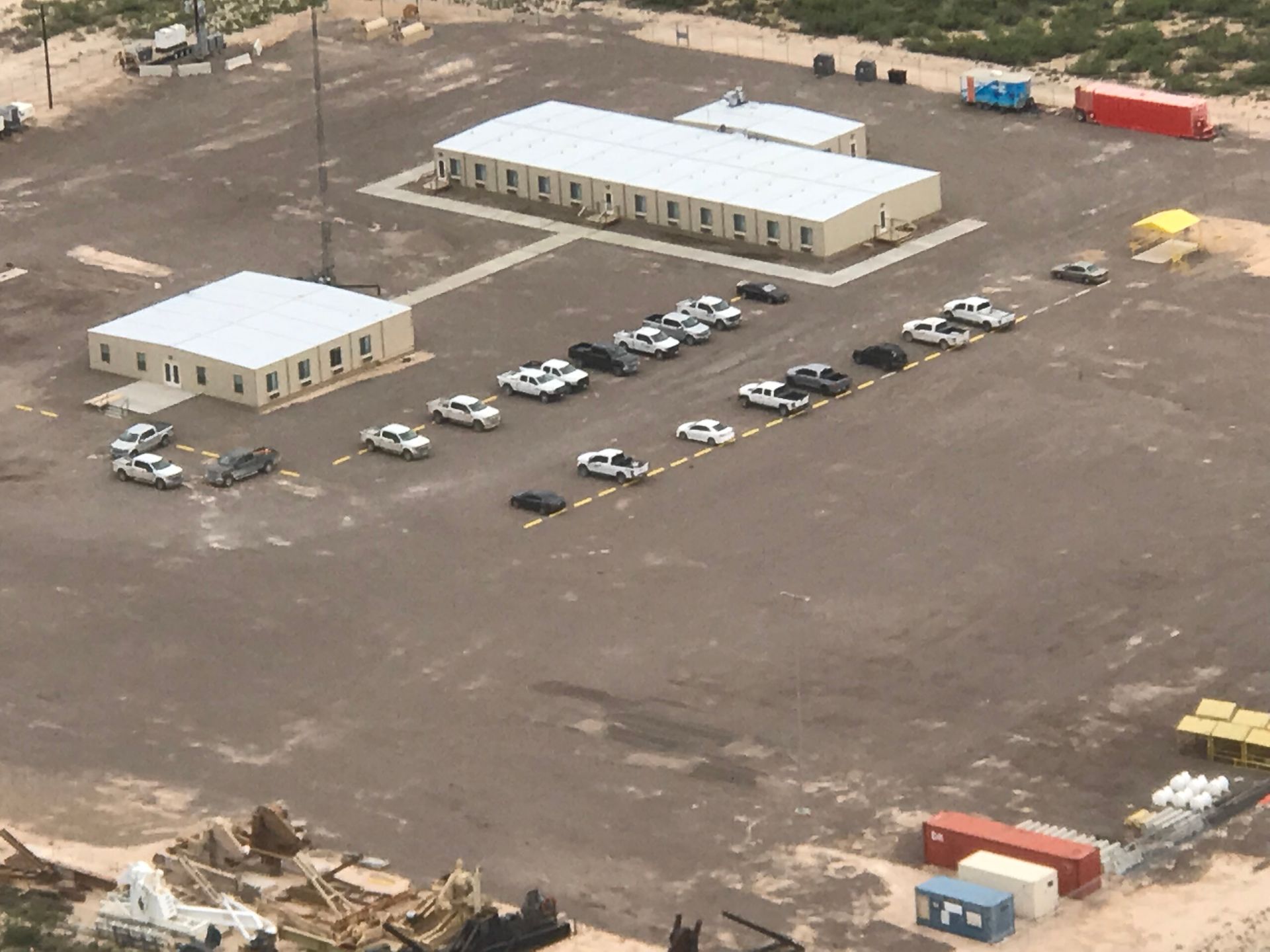 Aerial view of a dusty lot with two white buildings and several parked cars and trucks