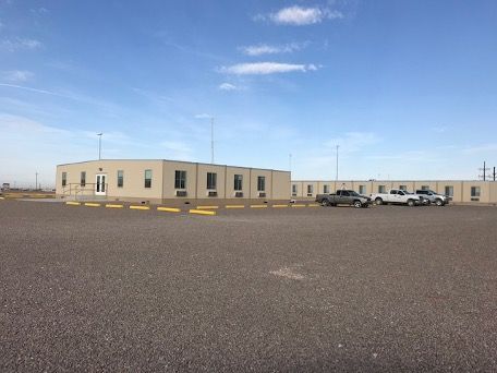 Low beige building in a large gravel lot under a blue sky, with a few parked cars.
