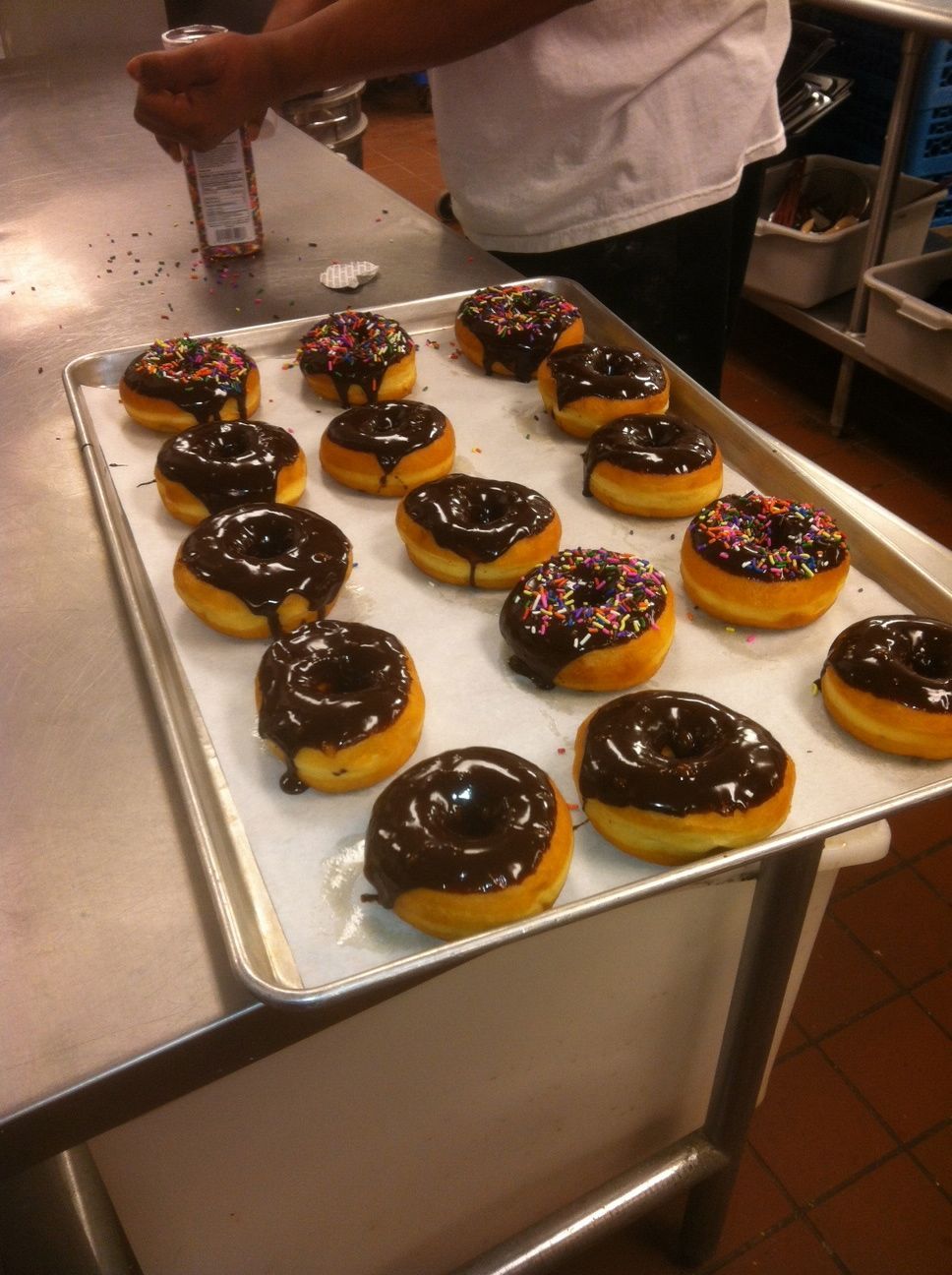 Chocolate-glazed donuts with colorful sprinkles arranged on a tray in a bakery kitchen.