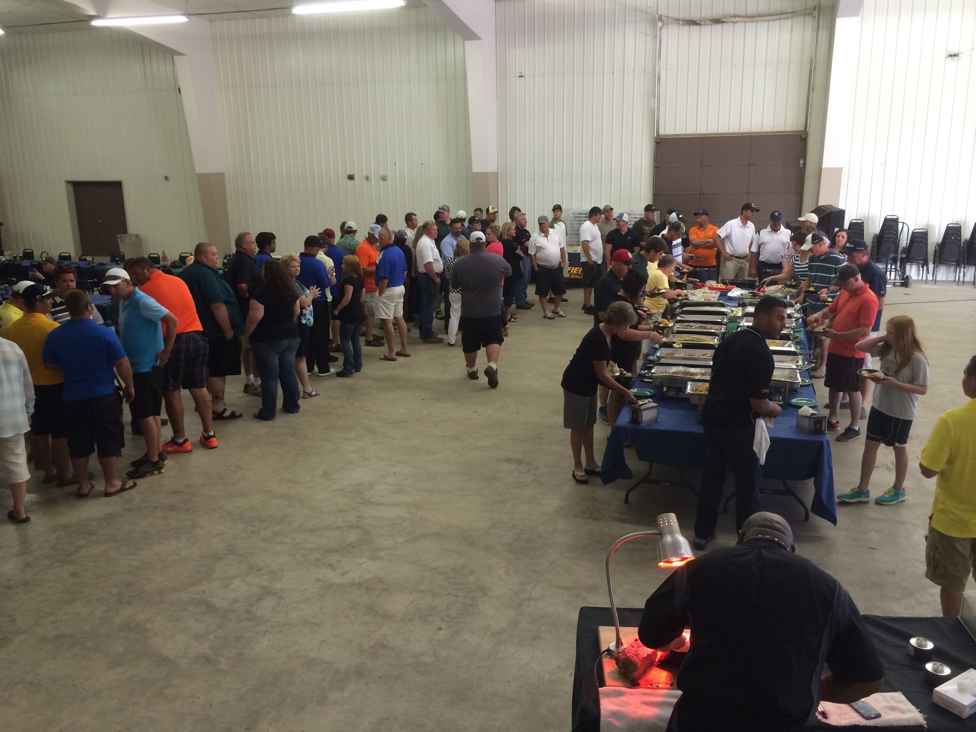 People lined up at buffet tables in a large indoor hall, with grills and trays in the foreground.