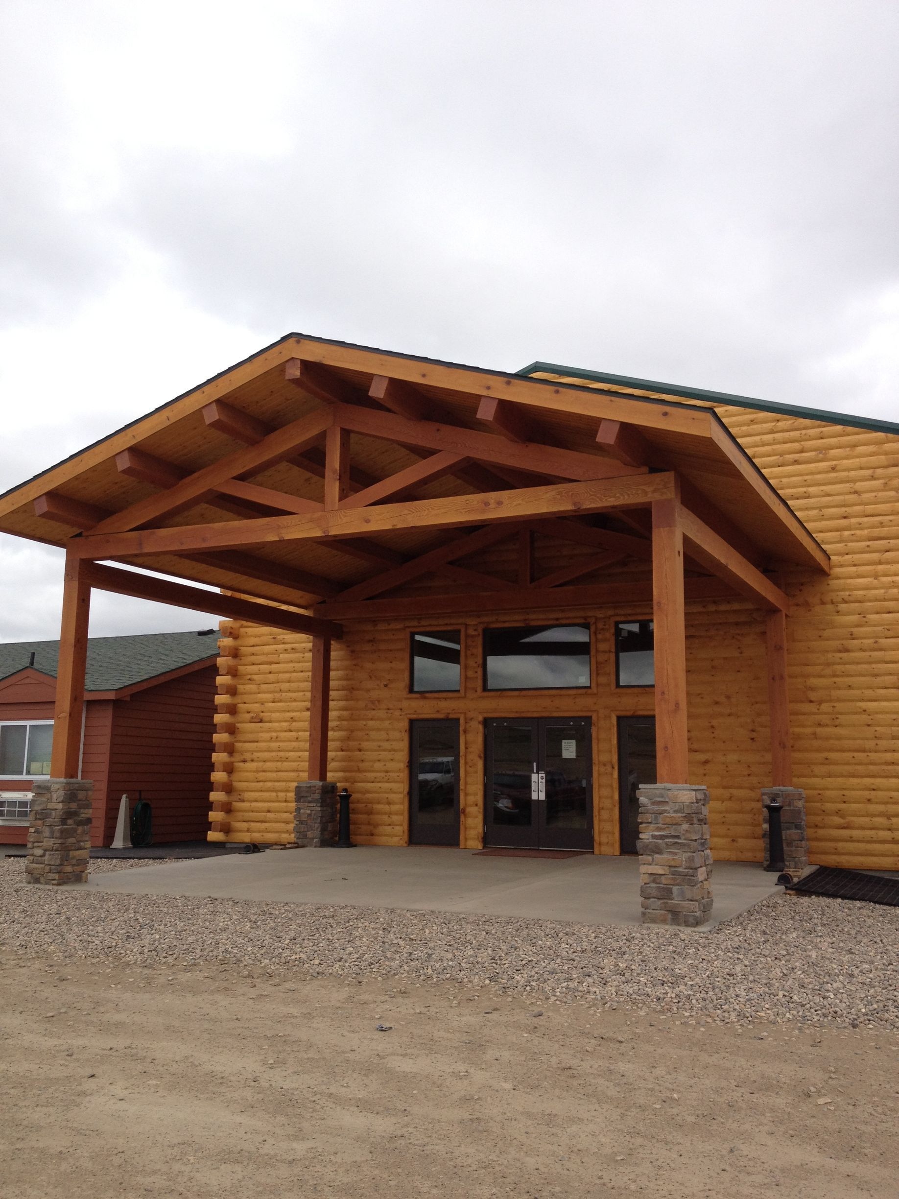 Wooden lodge entrance with covered porch, stone pillars, and gravel driveway under cloudy sky