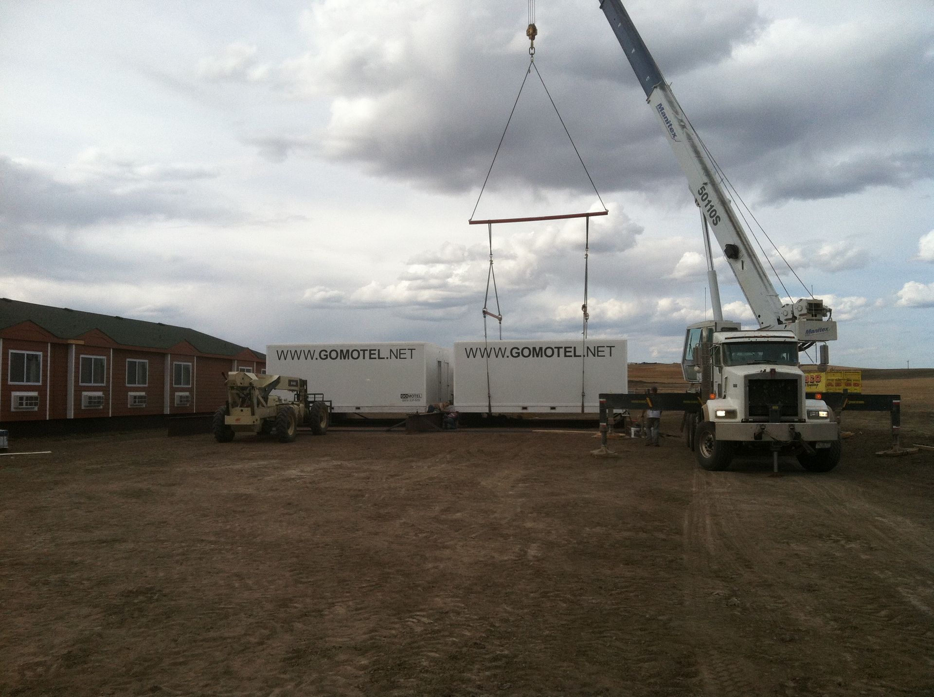 Crane lifting a white shipping container in a muddy yard beside a brick building under cloudy skies