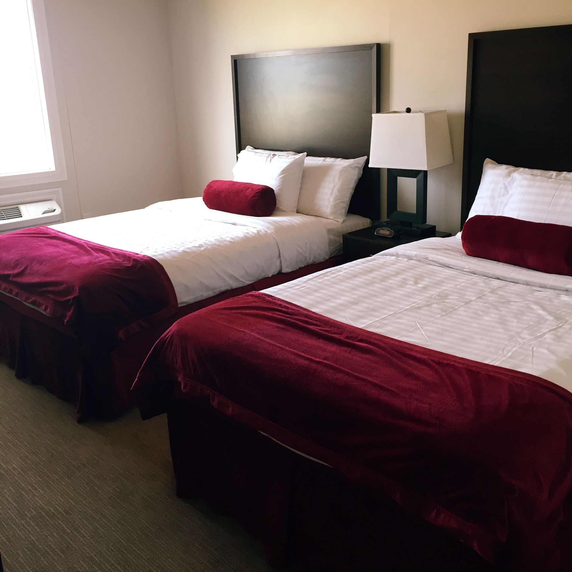 Hotel room with two twin beds, burgundy blankets, and a bedside lamp near a window.
