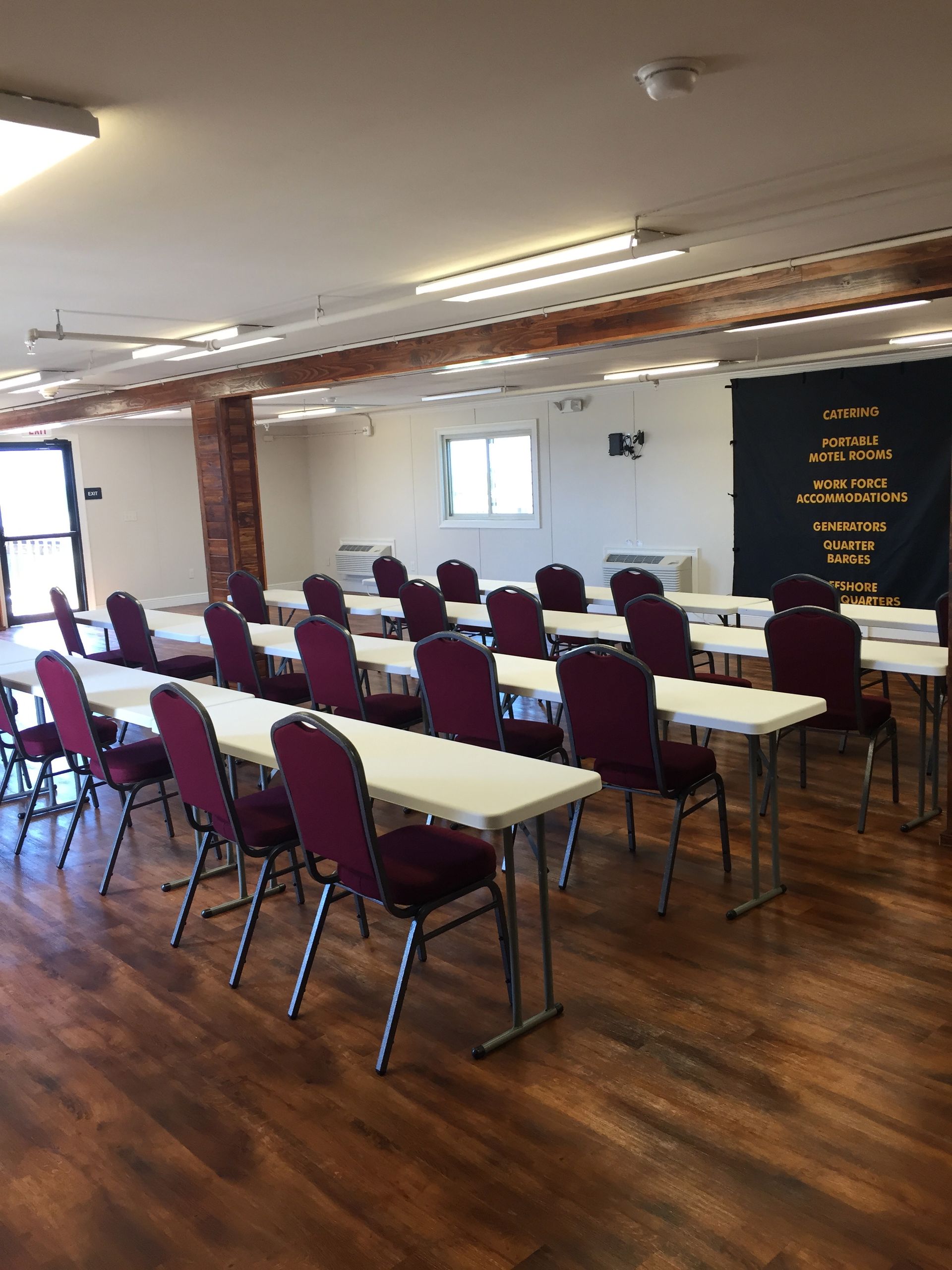 Empty classroom with rows of maroon chairs and tables on a wooden floor, lit by fluorescent ceiling lights