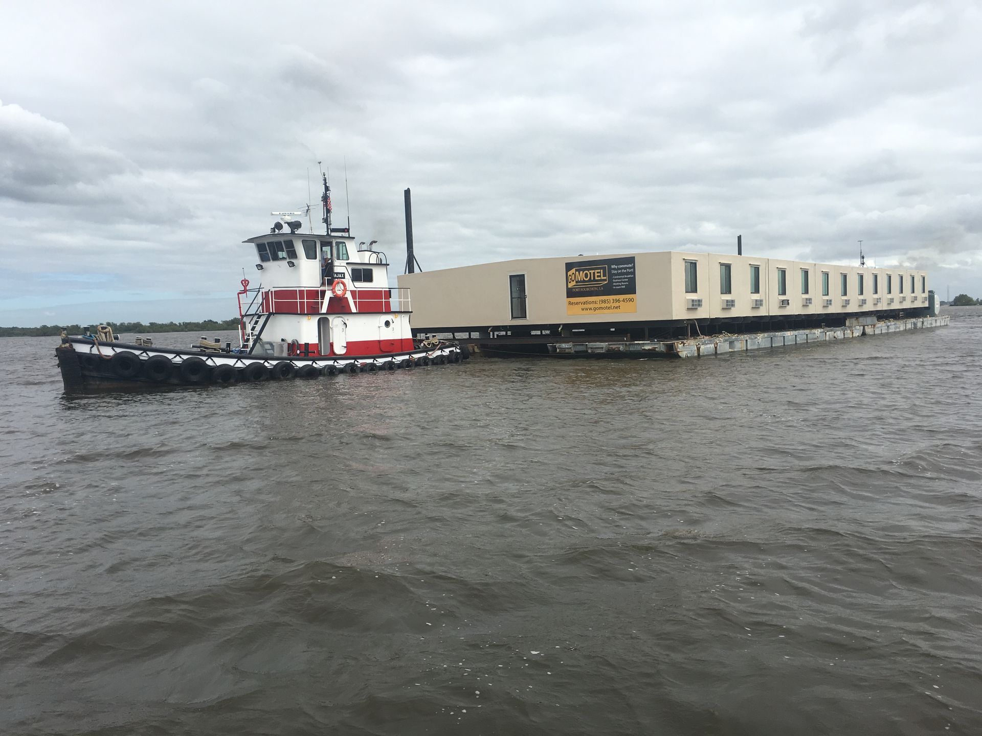 Red-and-white tugboat pushing a long beige barge on choppy water under a cloudy sky