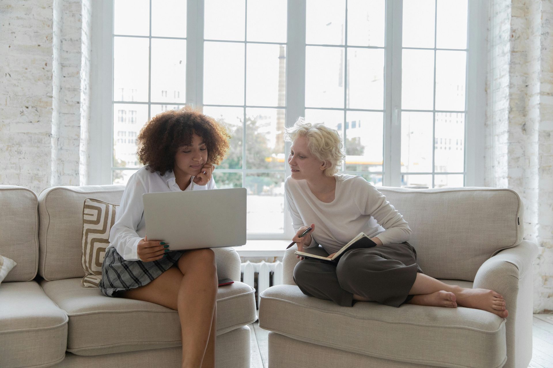 Two people on a couch, one using a laptop and the other holding a phone in a bright living room.
