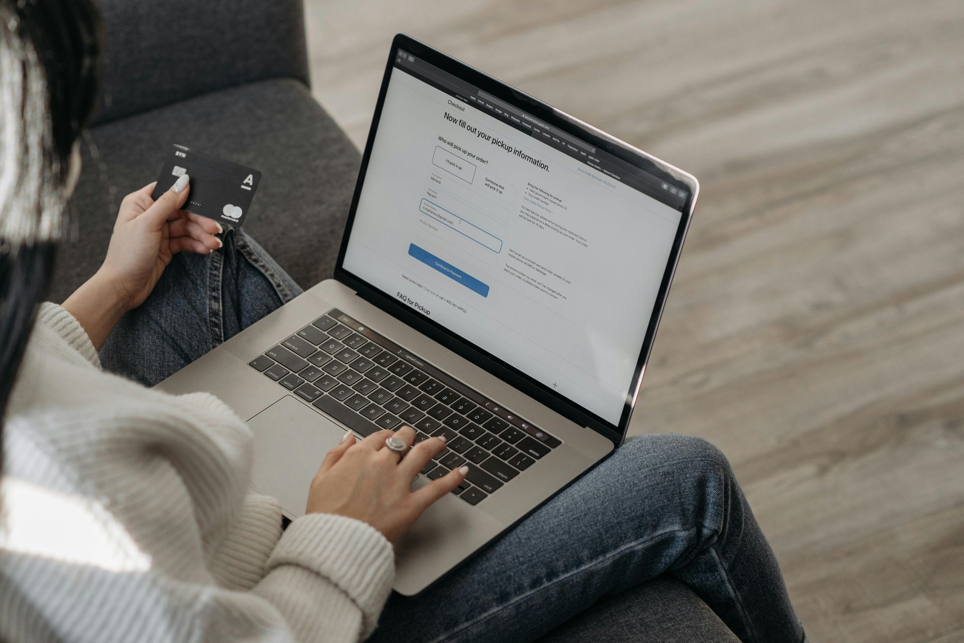 Woman in jeans on a couch, typing on a laptop and holding a credit card, possibly shopping online.