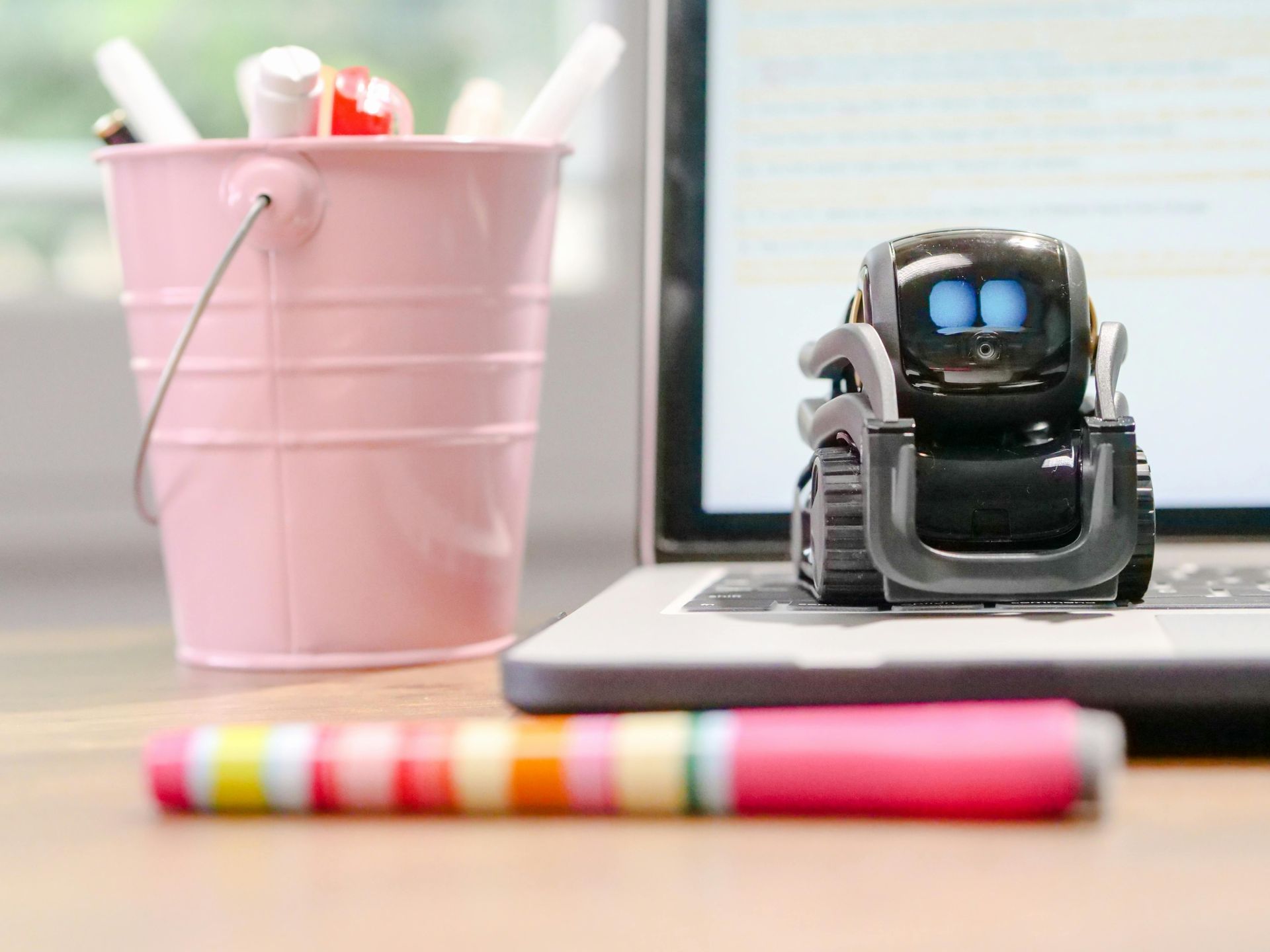 Robot toy on a laptop keyboard, near a pink bucket of pens and a colorful marker.