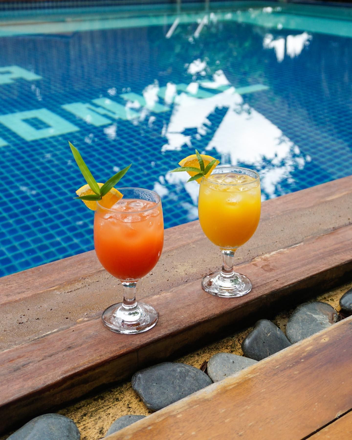 Two glasses of vibrant tropical cocktails with fruit garnishes placed on a wooden poolside deck next to a blue pool.