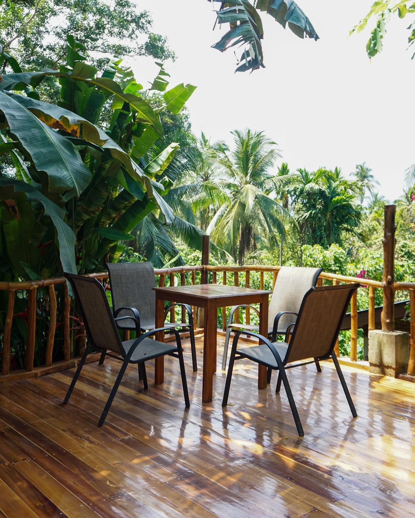A wooden balcony deck with a square table and four chairs surrounded by lush tropical trees and palms.
