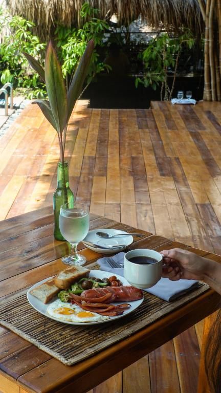 A breakfast plate with eggs, bacon, and toast sits on a wooden table outdoors, alongside a coffee mug and a glass of water.