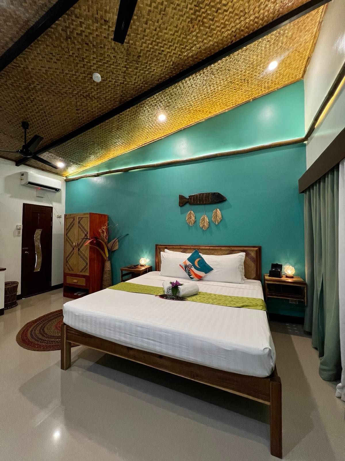 A hotel room with a wooden bed, teal wall, fish wall art, woven ceiling, and light green drapes.