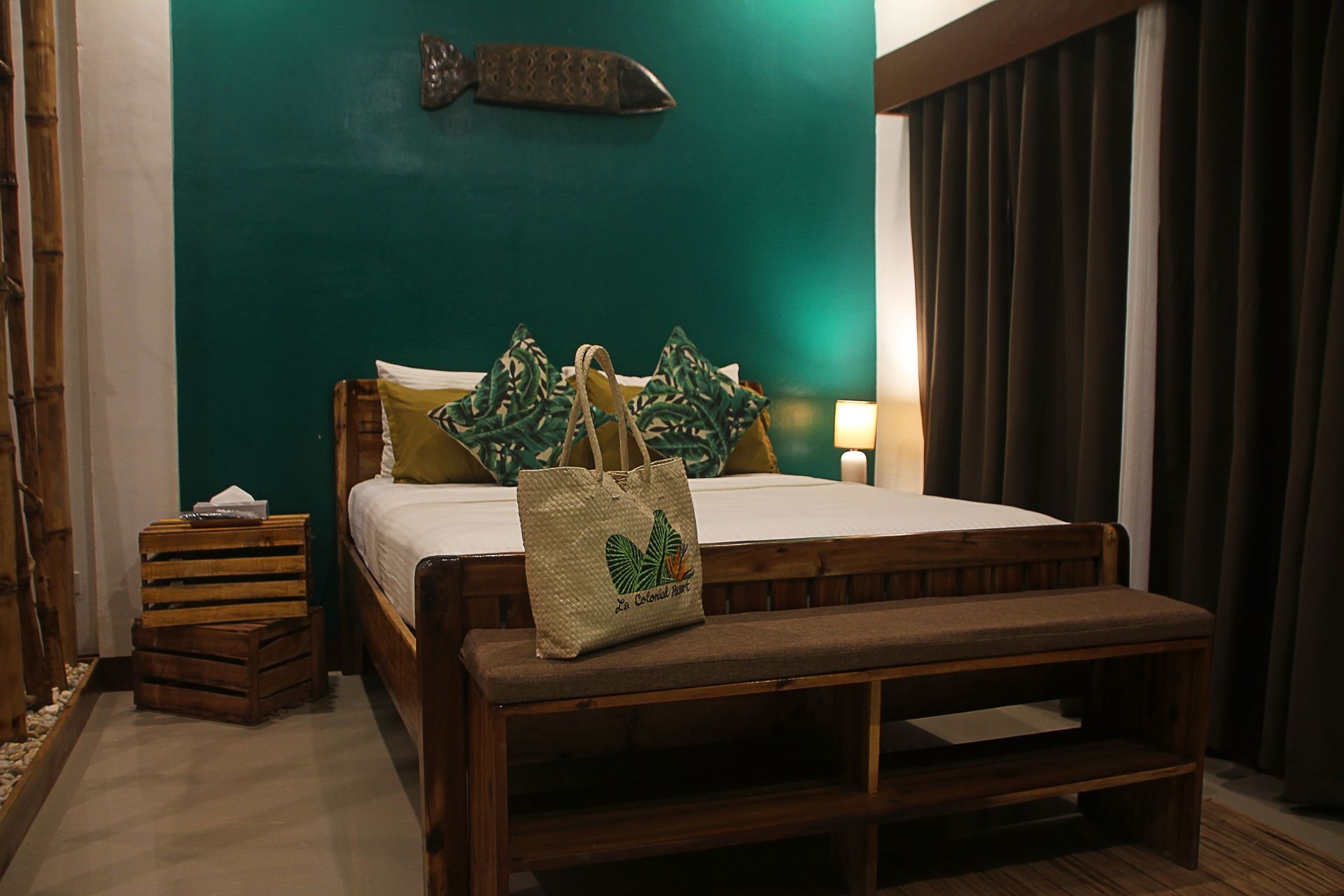 A bedroom featuring a bed with a white comforter, patterned pillows, a tote bag, and a wooden bench against a teal wall.