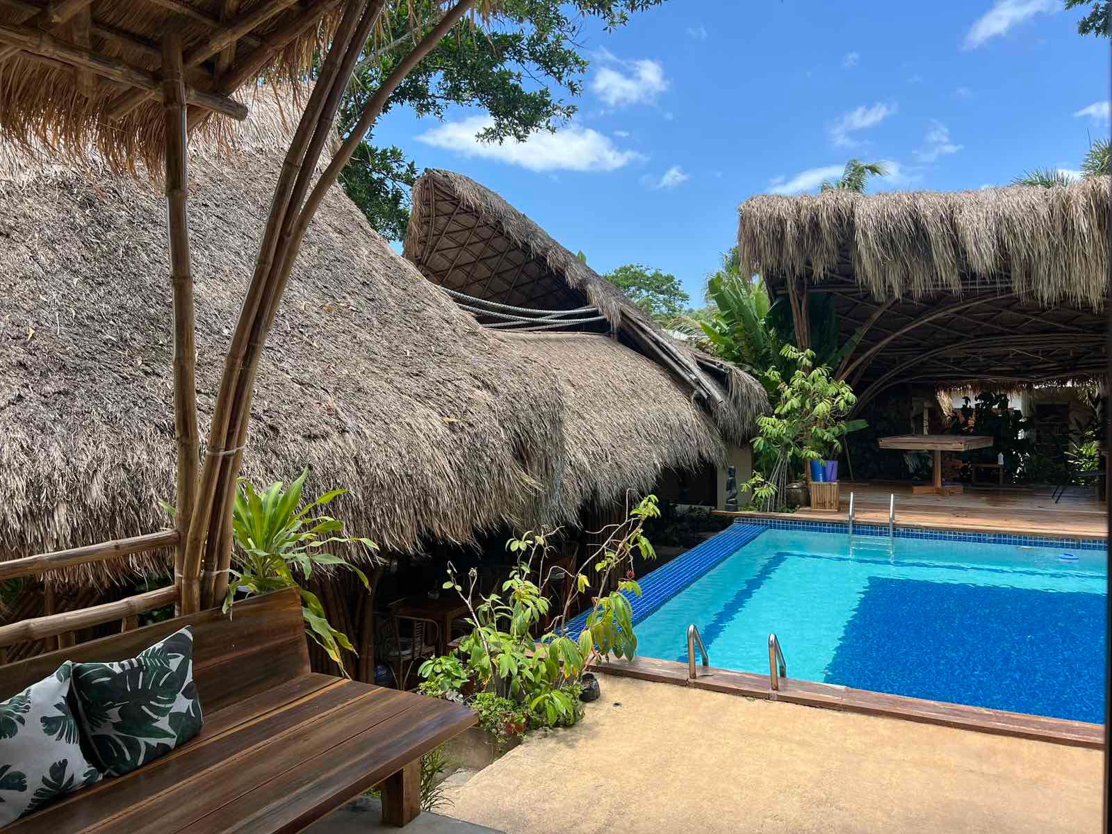 A swimming pool with blue water sits beside a thatched-roof building with a wooden bench in the foreground.