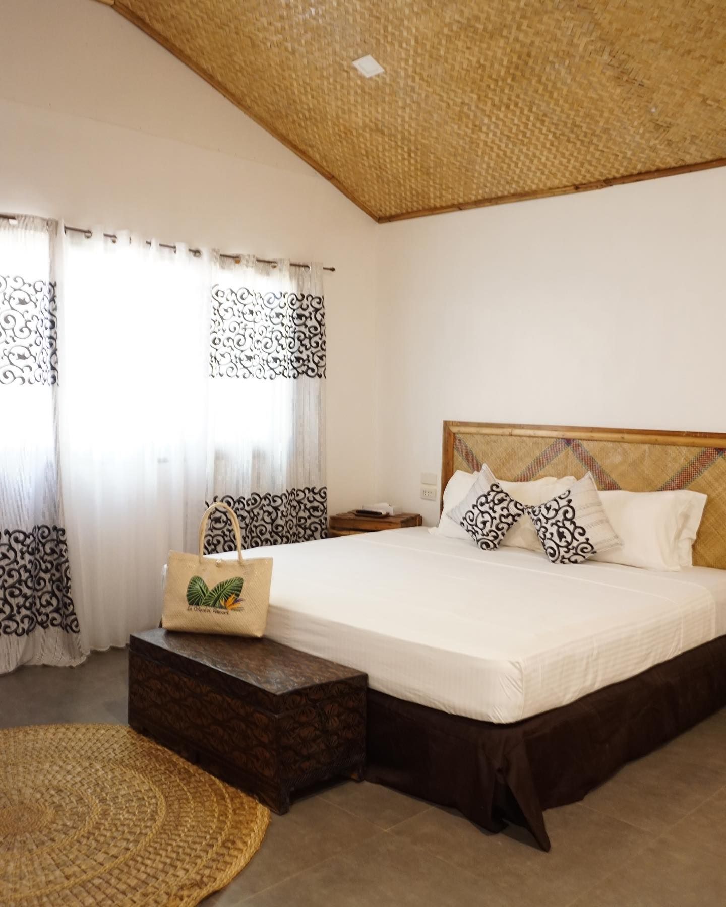 A bedroom with a bed, woven ceiling, decorative trunk, and patterned curtains, illuminated by natural light.
